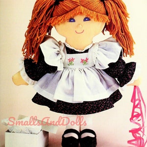 Peut inclure: Une poupée de chiffon faite à la main avec des cheveux en laine rouge en tresses, portant une robe noire et blanche avec un tablier blanc et des chaussures noires. La poupée tient un ruban rose et se tient à côté d'une boîte cadeau blanche avec du papier de soie.