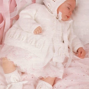 May include: A white knitted baby outfit with a matching hat and booties. The outfit features a delicate lace pattern and is tied with white ribbons.
