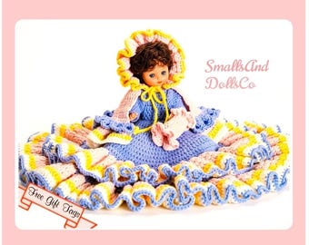 May include: A doll with brown hair wearing a blue crocheted dress with a wide, ruffled skirt in layers of pink, yellow, and blue. The doll has a matching bonnet and holds a small pink accessory. Text reads SmallsAnd DollsCo and Free Gift Tags.