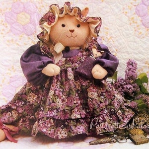 May include: A handmade plush cat doll wearing a purple dress with a floral print and a white lace collar. The doll is sitting on a white and pink quilt with a bouquet of purple flowers.