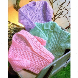 May include: Three knitted baby sweaters in pink, light green, and lavender with a cable knit pattern. The sweaters are folded and displayed in a wooden crate.