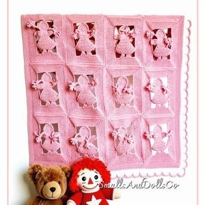 May include: A light pink crochet blanket featuring a grid of twelve rectangular panels, each containing a raised figure of a doll with pigtails. Below the blanket, a brown plush teddy bear and a doll with red yarn hair and a star shirt are visible.