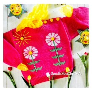 May include: A vibrant pink knitted sweater featuring daisy and sun motifs. The sweater has yellow detailing on the sleeves and neckline, with three yellow buttons. The text "SmallsAndDollsCo" is visible. Decorative sun and flower accents surround the sweater.