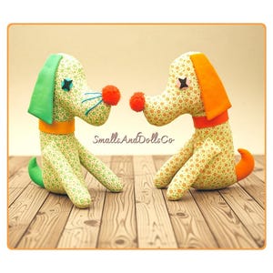 Puede incluir: Dos perros de peluche hechos de tela verde claro con un pequeño estampado floral. Cada perro presenta una nariz de pompón naranja brillante, ojos de botón y un collar de color sólido. Uno tiene orejas y cola verdes, mientras que el otro tiene detalles en naranja. El texto dice SmallsAndDollsCo.