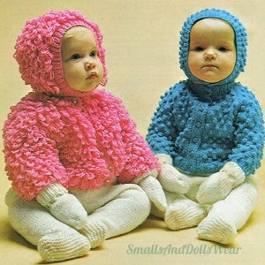 May include: Two babies wearing knitted hats and matching sweaters. The baby on the left is wearing a pink sweater and hat, while the baby on the right is wearing a blue sweater and hat. Both babies are wearing white pants.