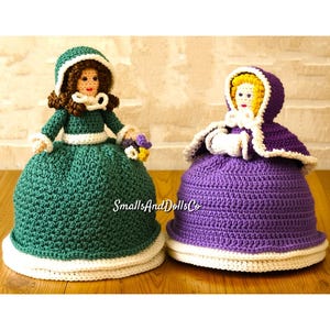 Puede incluir: Dos fundas de muñecas hechas a mano con ganchillo. Una es verde con ribete y capucha blancos, la otra es morada con ribete y capucha blancos. Ambas tienen caras de muñeca y están diseñadas para cubrir una muñeca.
