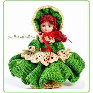 May include: A doll with red hair wearing a green and cream crocheted dress and a matching bonnet. The doll features a red ribbon tied at the neck and waist. The text SmallsAndDollsCo is visible on the left side of the doll against a white background.