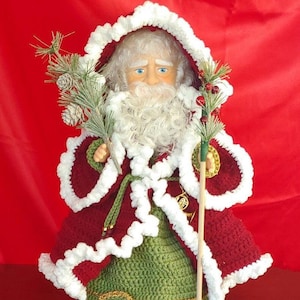 Può includere: Una bambola di Babbo Natale all'uncinetto con la barba bianca e un vestito rosso e verde. La bambola tiene un bastone di legno e sta in piedi su una base di legno.