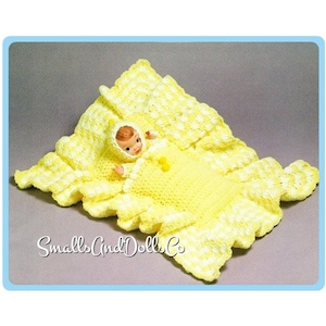 Könnte beinhalten: Eine gelb gehäkelte Babydecke mit passender Kapuze und einer kleinen Puppe darin. Die Decke hat einen gewellten Rand und ein weißes und gelbes Muster. Die Puppe trägt ein gelbes Outfit. Der Text "Smalls And Dolls Co" ist sichtbar.