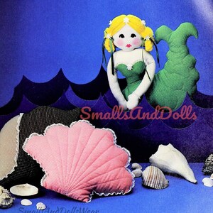 Puede incluir: Una muñeca de sirena verde con cabello amarillo y una almohada de concha rosa. La muñeca está sentada sobre un fondo azul con olas blancas. El texto "SmallsAndDolls" es visible en la imagen.
