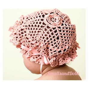 May include: A dusty pink crochet bonnet featuring an intricate openwork pattern, a central floral motif at the crown, and a ruffled edge. A matching satin ribbon is tied into a bow on the side. The text SmallsandDolls is visible in pink.