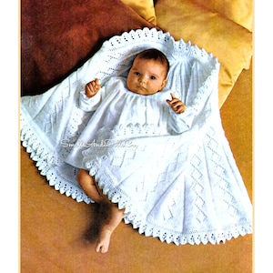 May include: A white crocheted baby blanket with a delicate lace border. The blanket is draped over a baby wearing a white dress.
