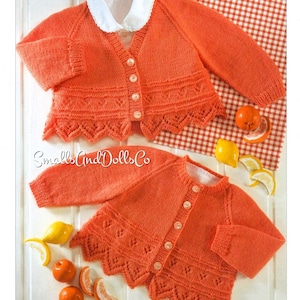 May include: Two orange knitted baby cardigans with button fastenings and scalloped edges. The cardigans are laid out on a white surface with lemons and oranges.