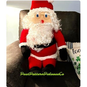 Puede incluir: Un muñeco de crochet de Santa Claus con un traje rojo, barba blanca y botas negras. El muñeco está sentado en un sofá marrón con una almohada blanca detrás.
