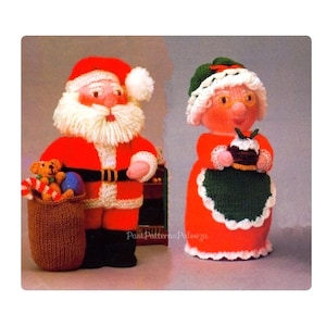 May include: Knitted Santa and Mrs. Claus dolls. Santa is wearing a red suit with white trim, a white beard, and a black belt. He is holding a bag of toys. Mrs. Claus is wearing a red dress with a green apron and a white hat. She is holding a Christmas pudding.
