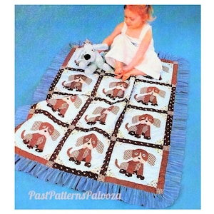 May include: A blue and brown patchwork baby quilt with a ruffled border. The quilt features a repeating pattern of brown dogs with white spots. A child is sitting on the quilt with a blue and white stuffed dog.