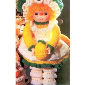 May include: Crocheted doll with yellow and white dress, green hat, and a yellow crocheted lemon in her hand. The doll is standing on a white and green crocheted base.