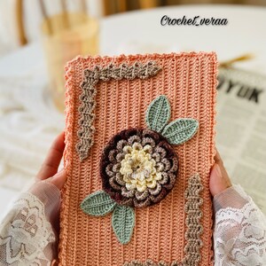 Puede incluir: Una bolsa de color melocotón hecha a crochet con un diseño de flor marrón, crema y verde. La bolsa tiene un borde de crochet con un borde festoneado.