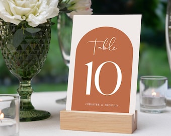 May include: A white card with a brown arch shape and the text "Table 10" in white. The card is on a wooden stand and the text "Christie & Richard" is at the bottom of the card.