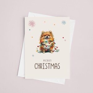 May include: A Christmas card featuring a cute brown and white Pomeranian dog wrapped in colorful Christmas lights. The card has a white background with a light brown border and the text "Merry Christmas" in black.