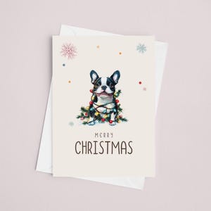 May include: A Christmas card featuring a black and white French bulldog wrapped in colorful Christmas lights. The card has a cream background with a light brown border and the text "Merry Christmas" in black.