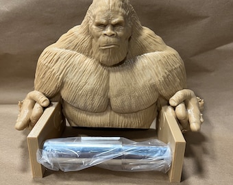 May include: A brown 3D printed toilet paper holder shaped like Bigfoot. The holder has a metal roll bar and is holding a roll of toilet paper.