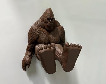 May include: A brown Bigfoot figurine with a textured fur-like appearance. The figurine is sitting with its legs extended, showcasing large feet. The figure has a stern expression and is likely a decorative item.
