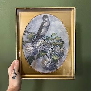 May include: Framed vintage print featuring a bird perched on a flowering branch. The artwork is set within an oval shape, surrounded by a gold-colored frame. The bird is depicted in shades of gray and black, with white flowers and green leaves.