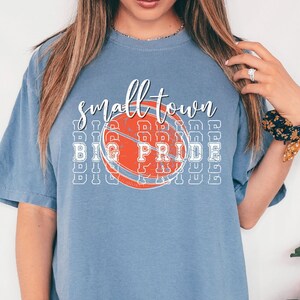 Puede incluir: Camiseta azul con la frase "small town big pride" en blanco y naranja, con un gr&aacute;fico de baloncesto naranja. La camiseta tiene un estilo casual y es c&oacute;moda.