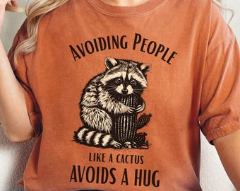 May include: Burnt orange t-shirt with a graphic of a raccoon hugging a cactus. The text reads "AVOIDING PEOPLE LIKE A CACTUS AVOIDS A HUG" in black. The shirt is worn with blue jeans.