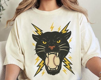 May include: Ivory-colored t-shirt featuring a black panther head with a baseball in its mouth, surrounded by yellow lightning bolts. The shirt is worn by a person sitting on a white couch. The word "IVORY" is printed in the upper right corner.