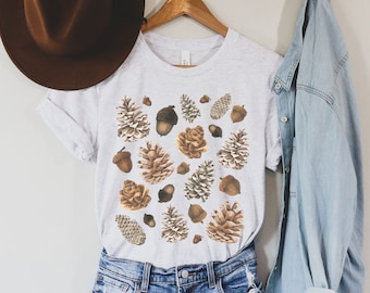 Puede incluir: Una camiseta gris claro con un estampado de bellotas y piñas en tonos marrones y beige. La camiseta se combina con pantalones cortos de mezclilla, un sombrero marrón y una camisa de mezclilla azul claro.