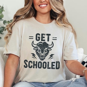 May include: A heather cement-colored t-shirt with the text "GET SCHOOLED" and a graphic of a bull's head. The shirt is worn with blue jeans. The model is sitting on a white couch with a black and white checkered blanket.