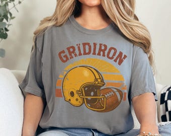 Könnte beinhalten: Graues T-Shirt mit einem Football-Design im Vintage-Stil. Die Grafik zeigt einen gelben Helm und einen braunen Football mit dem Wort "GRIDIRON" in einer Retro-Schriftart. Das Shirt ist ein bequemer, lässiger Stil.