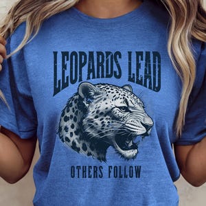 May include: A blue t-shirt with the text "LEOPARDS LEAD OTHERS FOLLOW" above and below a detailed illustration of a leopard's head. The leopard is depicted in black and white with its mouth open.