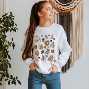 May include: White sweatshirt with a pattern of brown and beige pine cones and acorns.