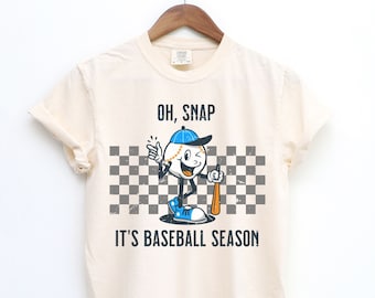 May include: A cream coloured t-shirt with a cartoon baseball character holding a bat. The text on the shirt reads "Oh, Snap It's Baseball Season".
