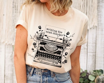 May include: A beige t-shirt with a black and white illustration of a vintage typewriter with the text "writing the next chapter" on a piece of paper in the typewriter.