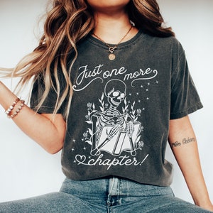 May include: A dark gray t-shirt with a white graphic of a skeleton reading a book. The graphic includes the text "Just one more chapter!" and floral accents.