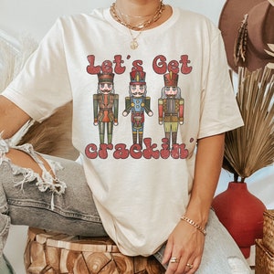 May include: A beige t-shirt featuring a vintage-style graphic of three nutcracker figurines and the text "Let's Get Crackin'" in red.