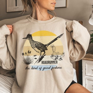 May include: A beige sweatshirt featuring a graphic print of a roadrunner bird in a desert landscape. The text "Roadrunner the bird of good fortune" is printed below the bird.