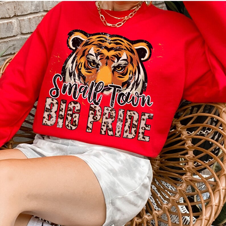Pu&ograve; includere: Felpa rossa con una grafica di tigre e il testo "Small Town Big Pride" in un font usurato. La grafica della tigre &egrave; di colore marrone e nero ed &egrave; circondata da un motivo a stampa leopardata.