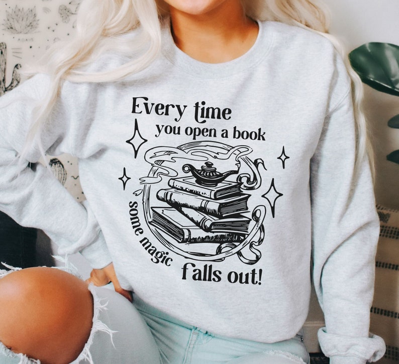 May include: A light gray sweatshirt with a black graphic design. The design features a stack of books, a genie lamp, and the text "Every time you open a book some magic falls out!" The design is surrounded by stars and swirls.