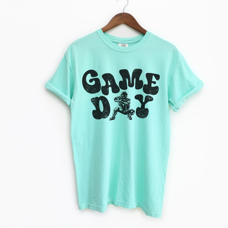 May include: A light blue t-shirt with a black graphic print of a person playing a sport. The graphic text reads "GAME DAY" in a retro font.