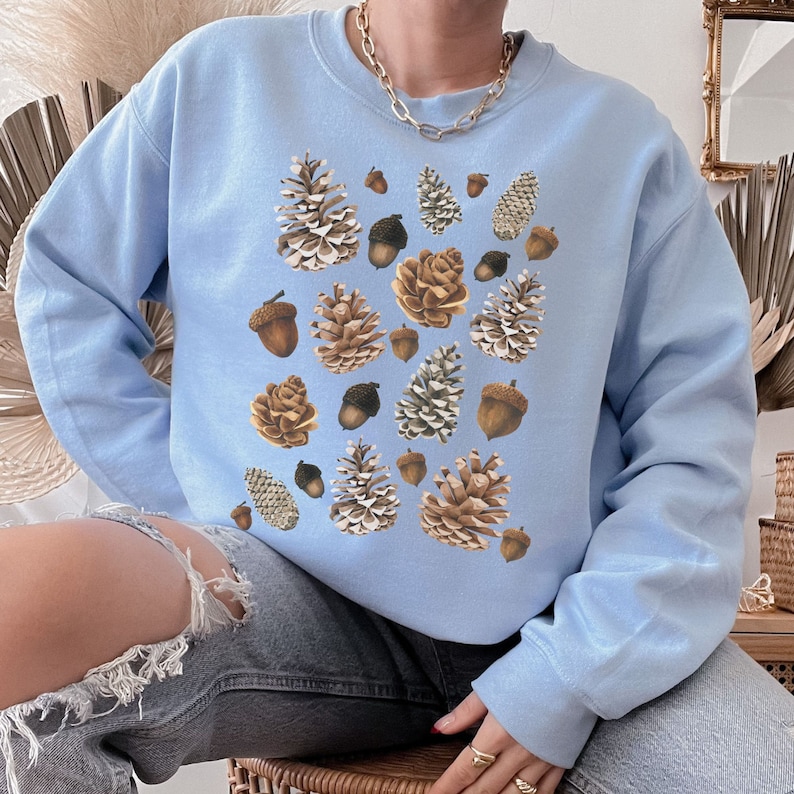 May include: A light blue sweatshirt with a pattern of brown pine cones and acorns. The sweatshirt has a crew neck and long sleeves.