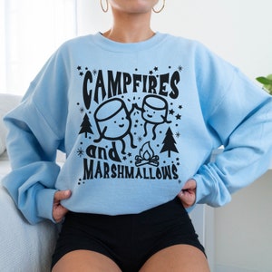 Puede incluir: Una sudadera de color azul claro con un dise&ntilde;o gr&aacute;fico negro de dos malvaviscos tom&aacute;ndose de la mano frente a una fogata con el texto "Campfires and Marshmallows".