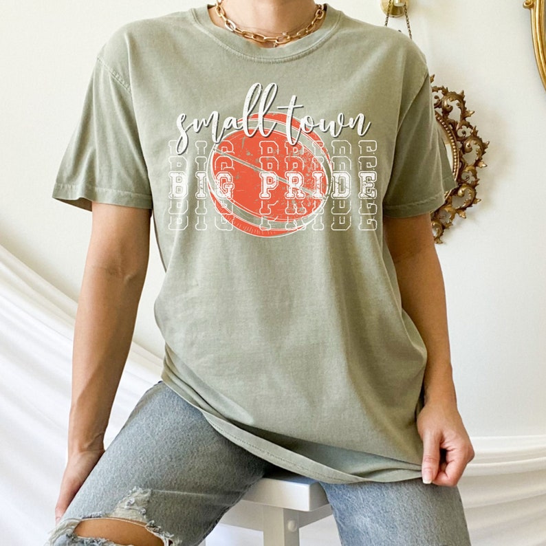 Puede incluir: Camiseta verde salvia con las palabras "Small Town Big Pride" en escritura blanca y letras de bloque. Un gr&aacute;fico de baloncesto naranja desgastado est&aacute; centrado en la camiseta. La camiseta es de un estilo c&oacute;modo e informal.