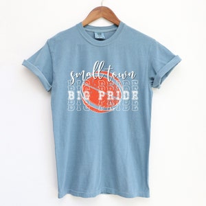 Puede incluir: Camiseta azul claro con las palabras "small town" sobre un gr&aacute;fico de baloncesto que dice "BIG PRIDE". La camiseta est&aacute; colgada en una percha de madera sobre un fondo blanco. La camiseta tiene las mangas enrolladas.