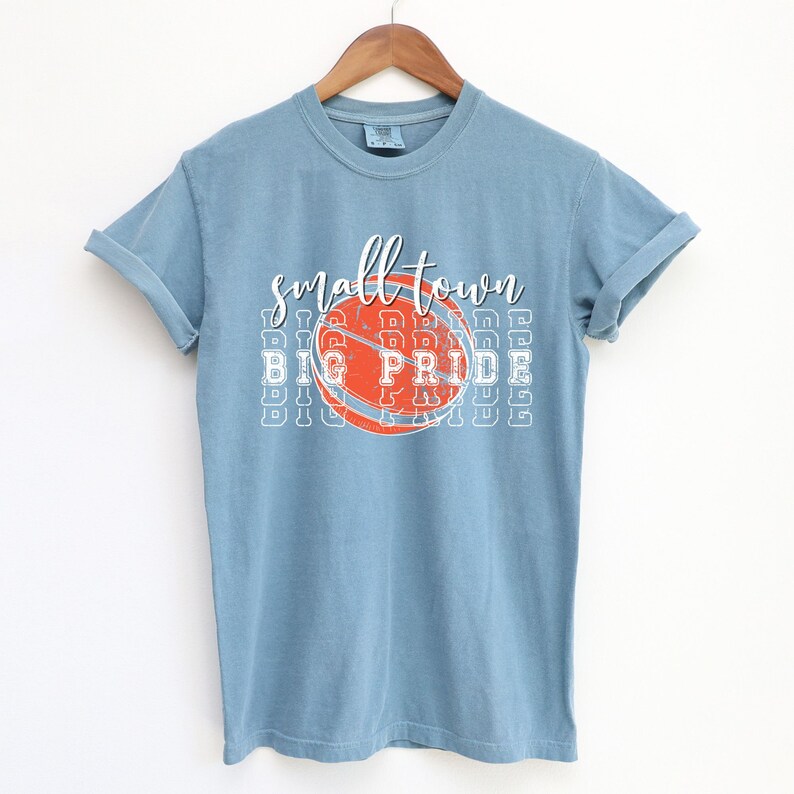 Puede incluir: Camiseta azul claro con las palabras "small town" sobre un gr&aacute;fico de baloncesto que dice "BIG PRIDE". La camiseta est&aacute; colgada en una percha de madera sobre un fondo blanco. La camiseta tiene las mangas enrolladas.