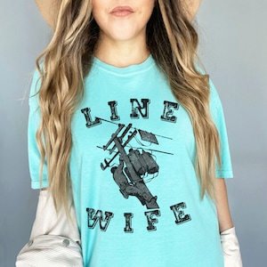 May include: A light blue t-shirt with a graphic of a lineman working on a power line. The text "LINE WIFE" is printed in black letters on the shirt.